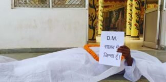 DM साहब मैं जिंदा हूँ…बस्ती में कफन ओढ़कर डीएम ऑफिस के बाहर लेट गया बुजुर्ग, मचा बवाल